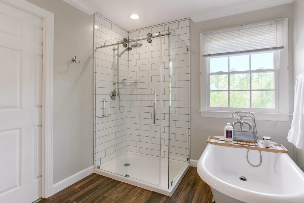 Bright bathroom with glass shower, white walls, wood floor, and a clawfoot tub by a window