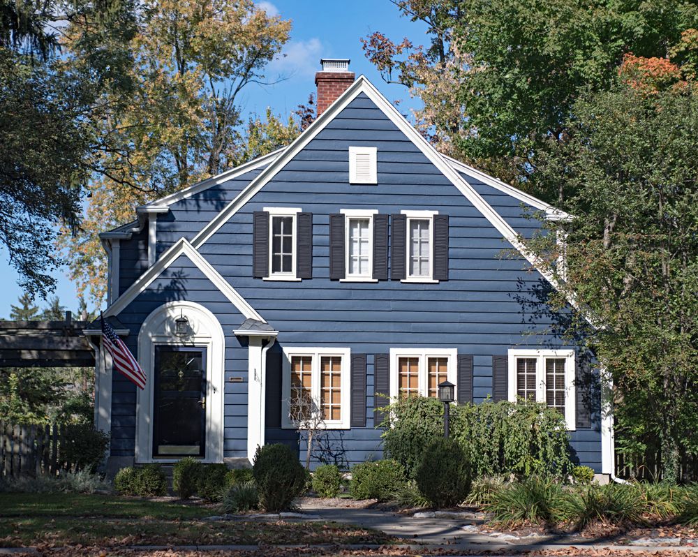 Blue house with white trim and black shutters, trees in background, American flag by the door.