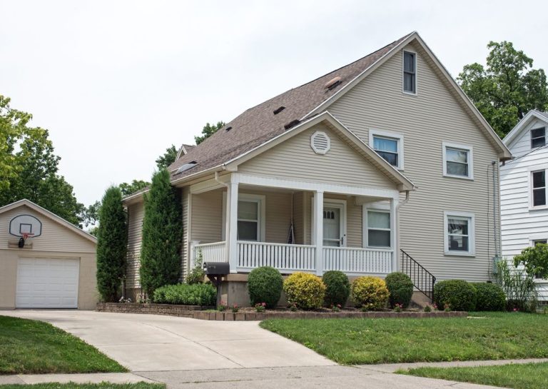 Beige two-story house with a porch and attached garage, on a green lawn.