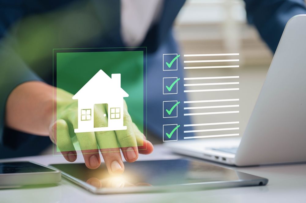 Person using a tablet with a house icon and checklist overlaid, beside a laptop.