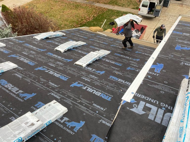 Roofer using a nail gun to install gray shingles on a rooftop. Worker wearing safety vest and gloves.