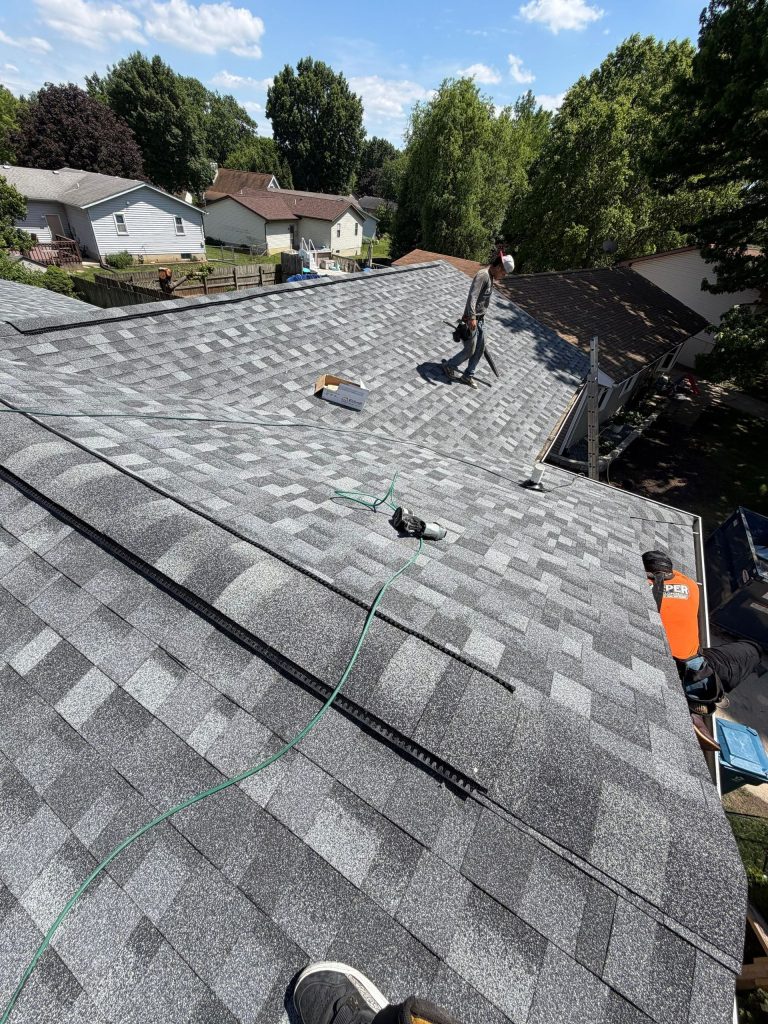 Roofers installing gray shingles on a steep residential roof under a blue sky