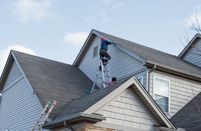 Two workers on a house roof: one on a ladder, the other near a window, both wearing hats.