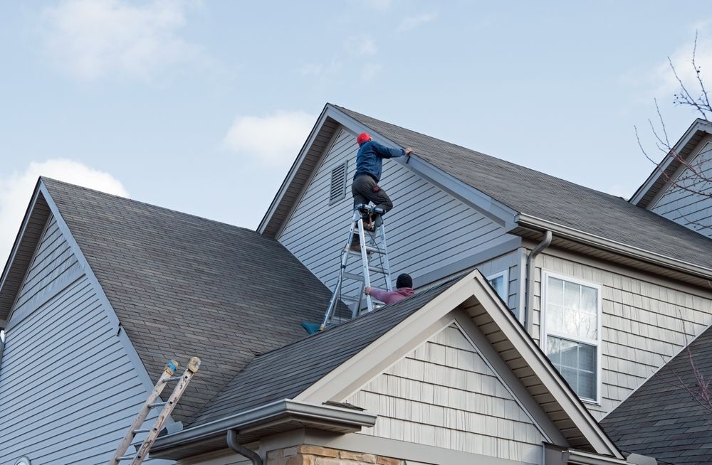 Two workers on a house roof: one on a ladder, the other near a window, both wearing hats.