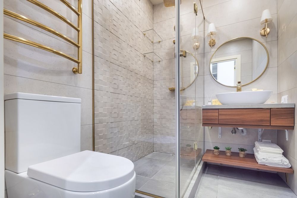 Modern bathroom with a white toilet, glass-enclosed shower, wood vanity with a circular mirror, and gold fixtures.