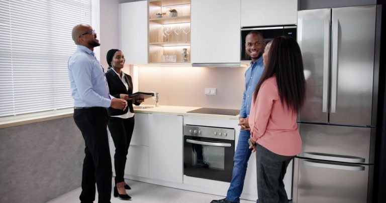A couple viewing a kitchen with a realtor. Stainless steel appliances, white cabinets, and overhead lighting.