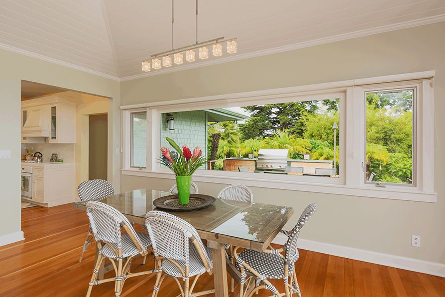 Dining room with glass table, white wicker chairs, and a large window overlooking a yard.