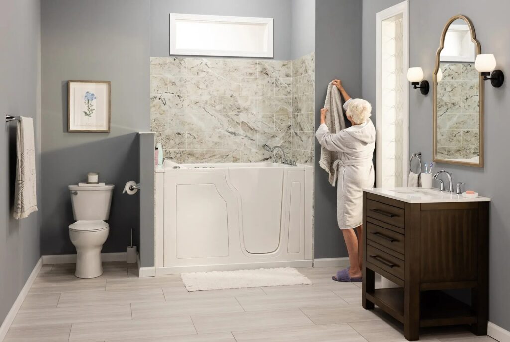 A bathroom with a white walk-in tub set in a marble-look wall surround, beside a dark wood vanity, with a woman in a robe hanging a towel.