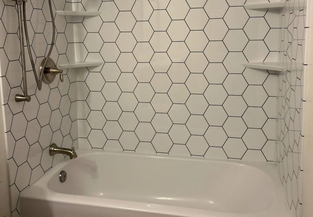 A white tub with a white hexagon tile-look wall surround, corner shelves and a brushed nickel handheld shower.