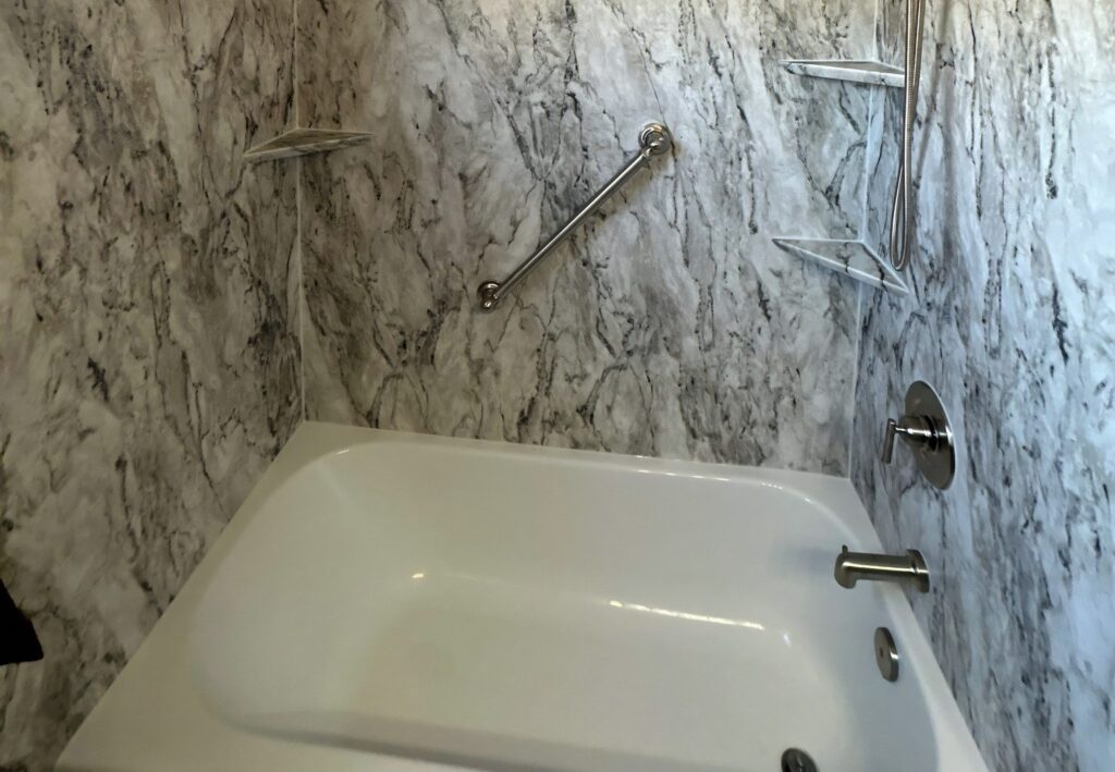 A white tub with a gray and white marble-look wall surround, corner shelves, a grab bar and a brushed nickel faucet.